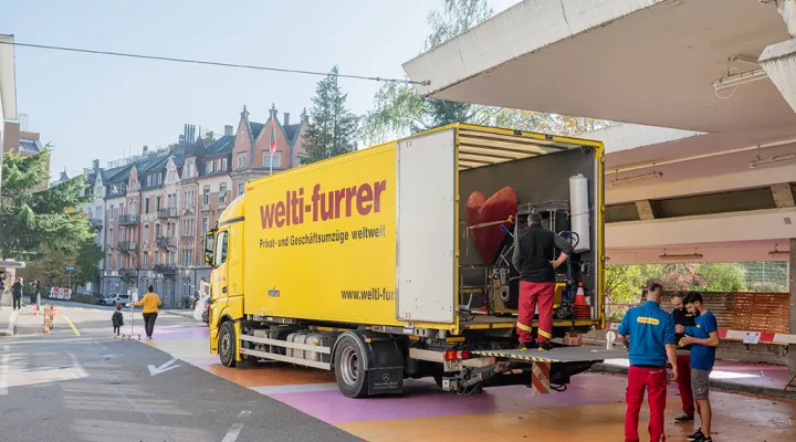 Drei Männer verladen Güter in einem gelben Lastwagen mit der Aufschrift "Welti-Furrer"