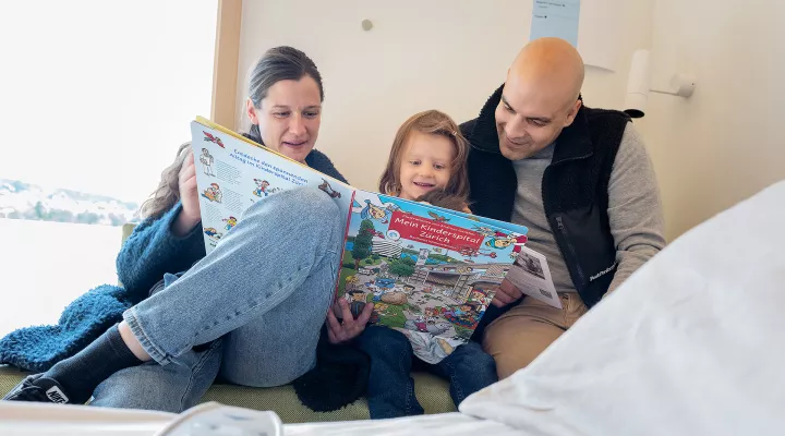 Eine Frau, ein Kind und ein Mann lesen ein Buch. Sie sitzen auf einer Bank neben einem Spitalbett.