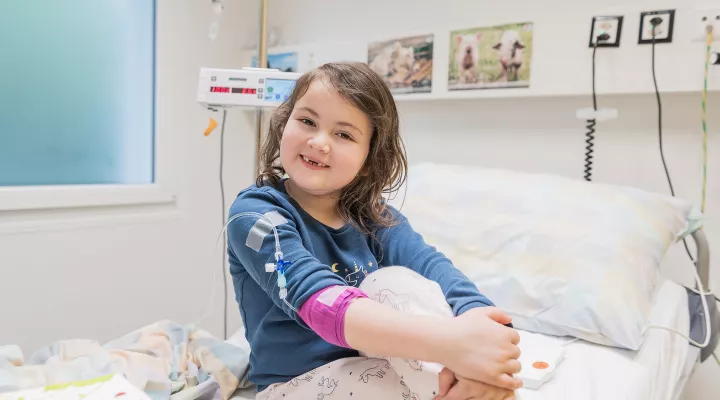 Eine Rheumapatientin sitzt im Schlafanzug auf einem Spitalbett. Sie hat ihre Beine überkreuzt und hat ihre rechte Hand auf die linke Hand abgelegt.