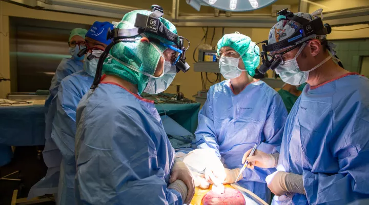 Ärzte in blauer Uniform, mit Masken und grünen Hauben stehen um einen OP-Tisch, darauf ist eine offene Gebärmutter zu erkennen.