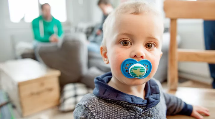 Themenbild zu Studie von Entwicklungspädiatrie, kleiner Junge mit grossem Schnuller blickt direkt in die Kamera, im Hintergrund unscharf sitzen die Eltern auf dem Sofa