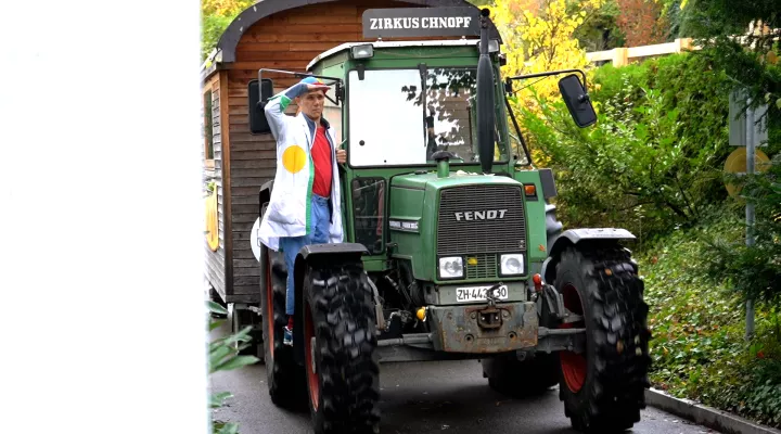 Ein grüner Traktor mit der Aufschrift "Zirkus Chnopf" zieht einen Zirkuswagen, ein Spitalclown hält sich an der Wagentür fest und blickt voraus