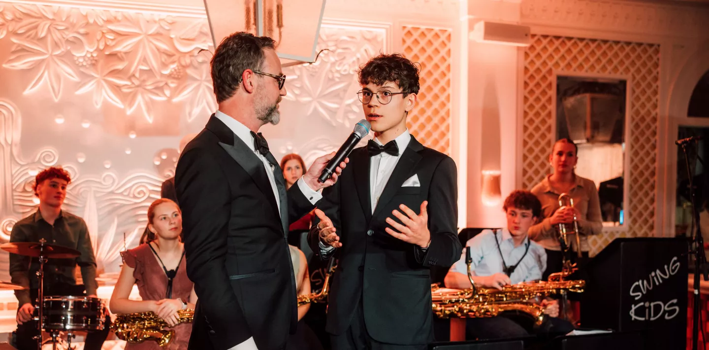 Zwei Männer in Anzügen stehen auf der Bühne. Einer hält das Mikrofon, der andere spricht hinein und gestikuliert dabei. Im Hintergrund sitzt eine Band mit ihren Instrumenten. 