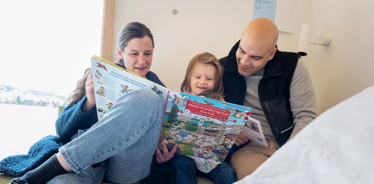 Eine Frau, ein Kind und ein Mann lesen ein Buch. Sie sitzen auf einer Bank neben einem Spitalbett.