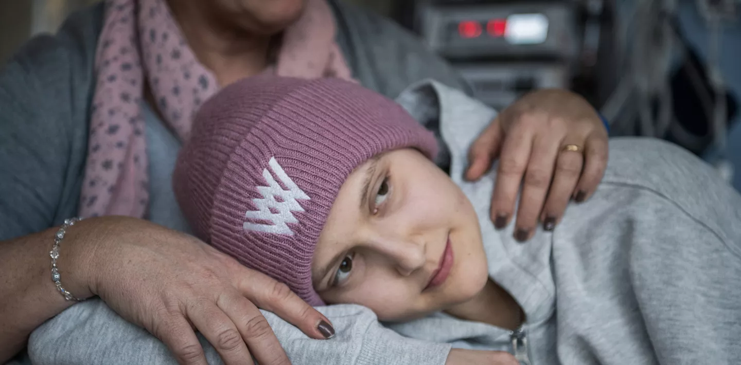 Eine Onkologiepatientin trägt eine Mütze und einen Pullover. Sie liegt auf dem Bett. Im Hintergrund sieht man den Oberkörper ihrer Mutter. 