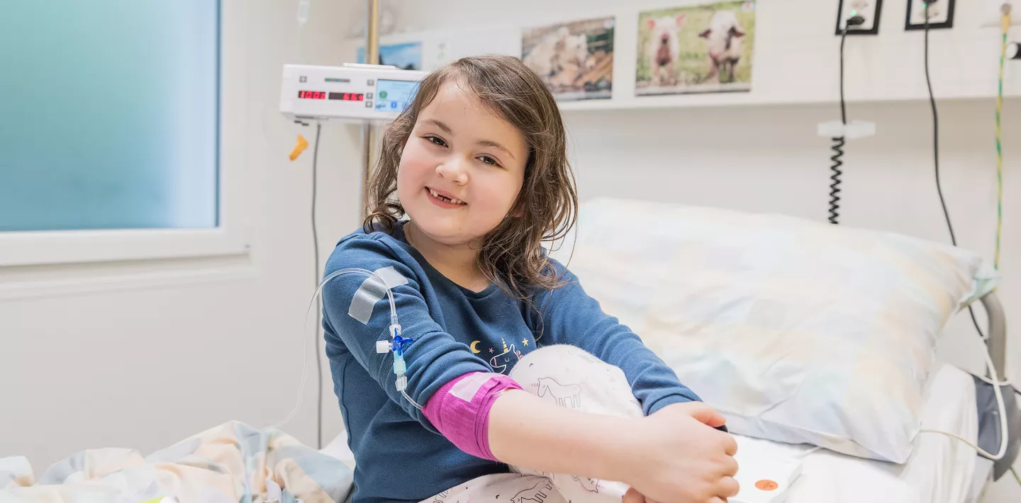 Eine Rheumapatientin sitzt im Schlafanzug auf einem Spitalbett. Sie hat ihre Beine überkreuzt und hat ihre rechte Hand auf die linke Hand abgelegt.