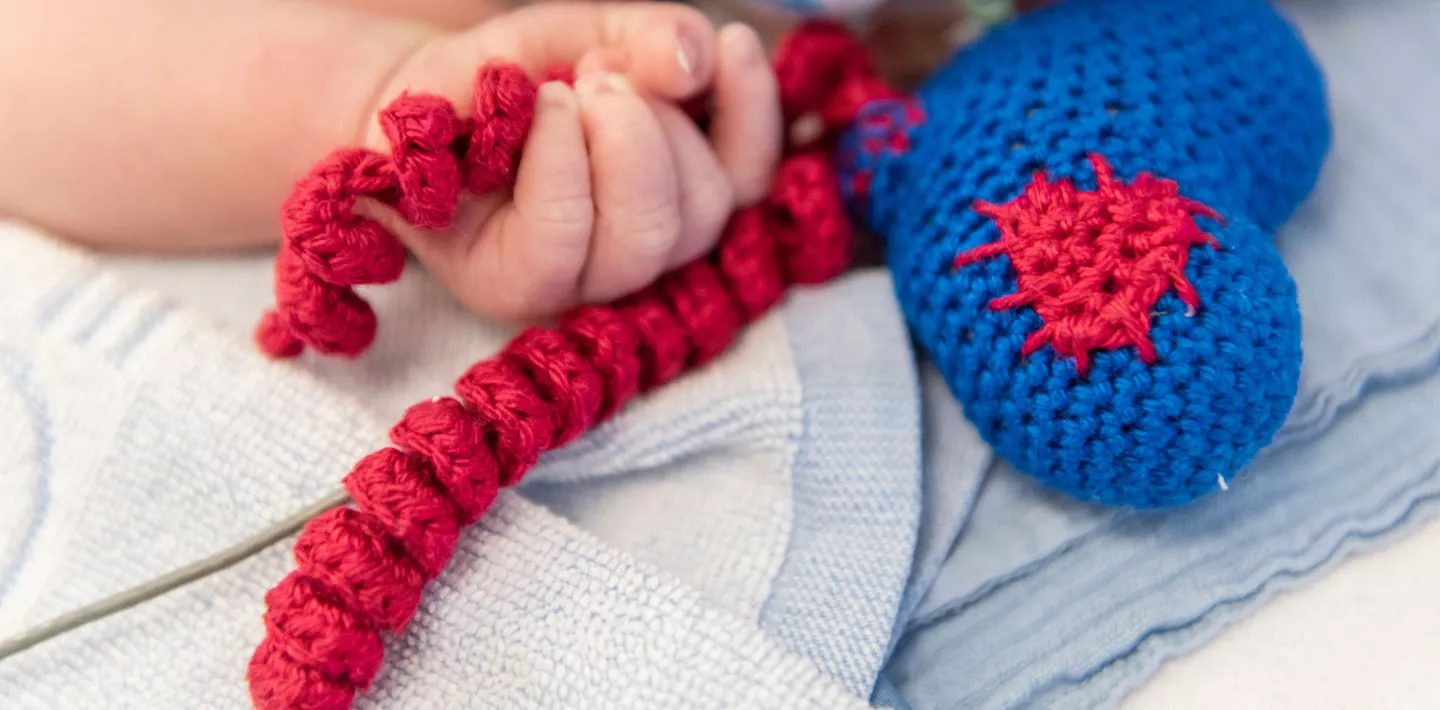 Ein neugeborenes Kind auf der Neonatologie hält ein gestricktes Herz in er Hand um den Greifreflex zu unterstützen