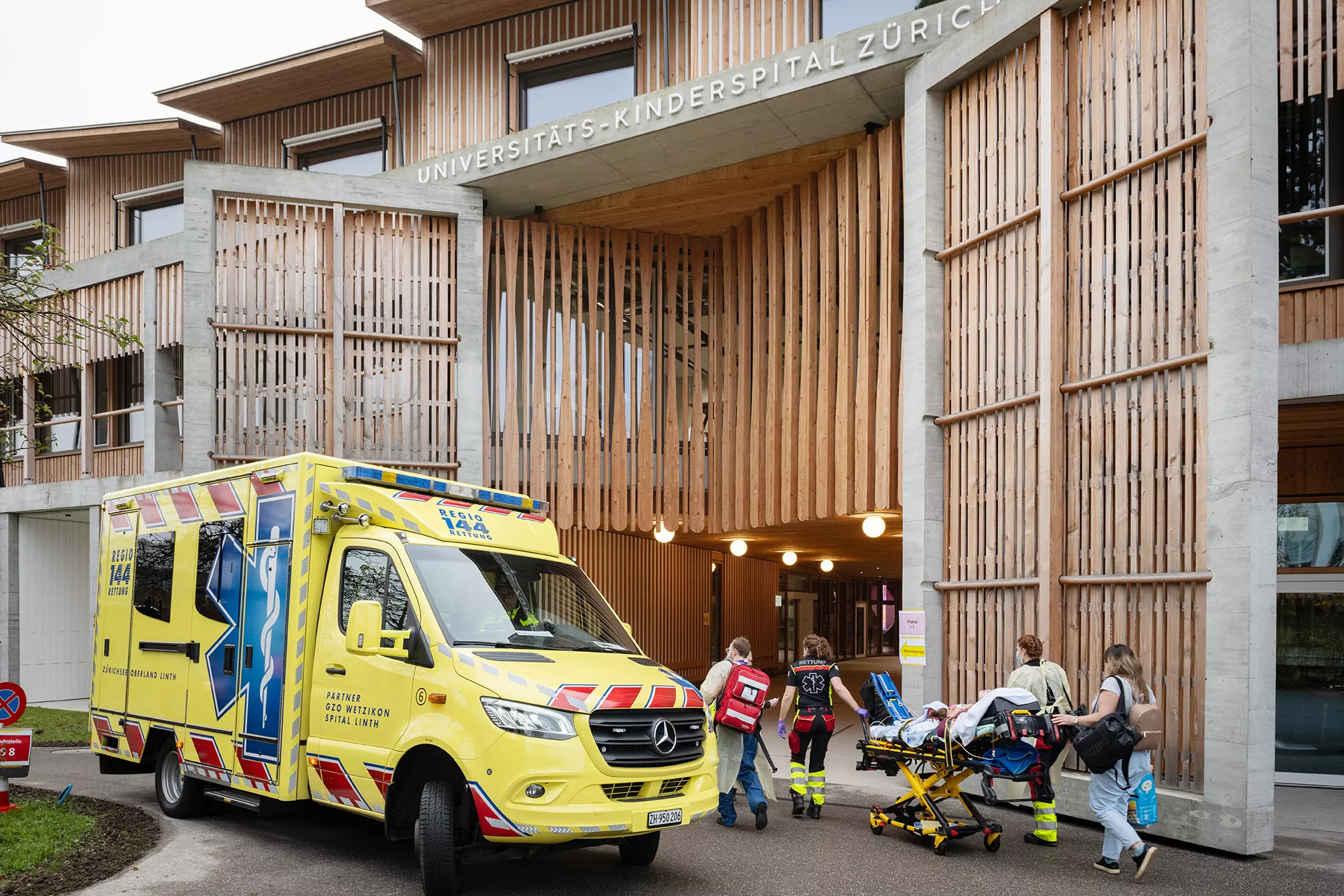 Ein gelber Ambulanzwagen steht vor dem Kinderspital Zürich, medizinische Fachpersonen transportieren ein Kind auf einer Liege