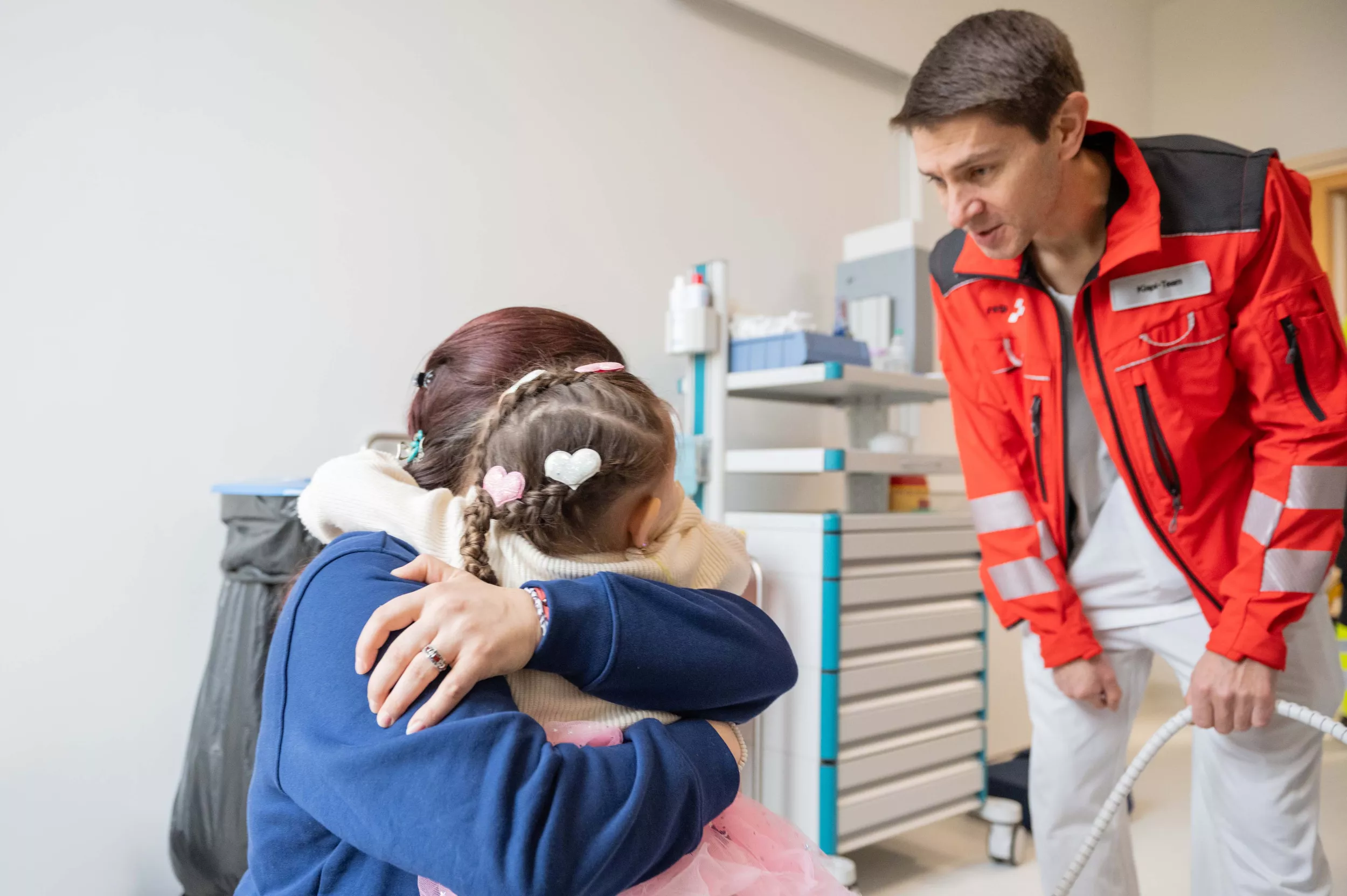 Eine Mutter umarmt ihre Tochter, daneben steht ein Mann mit einer roten Jacke und der Aufschrift "Kispi-Team"