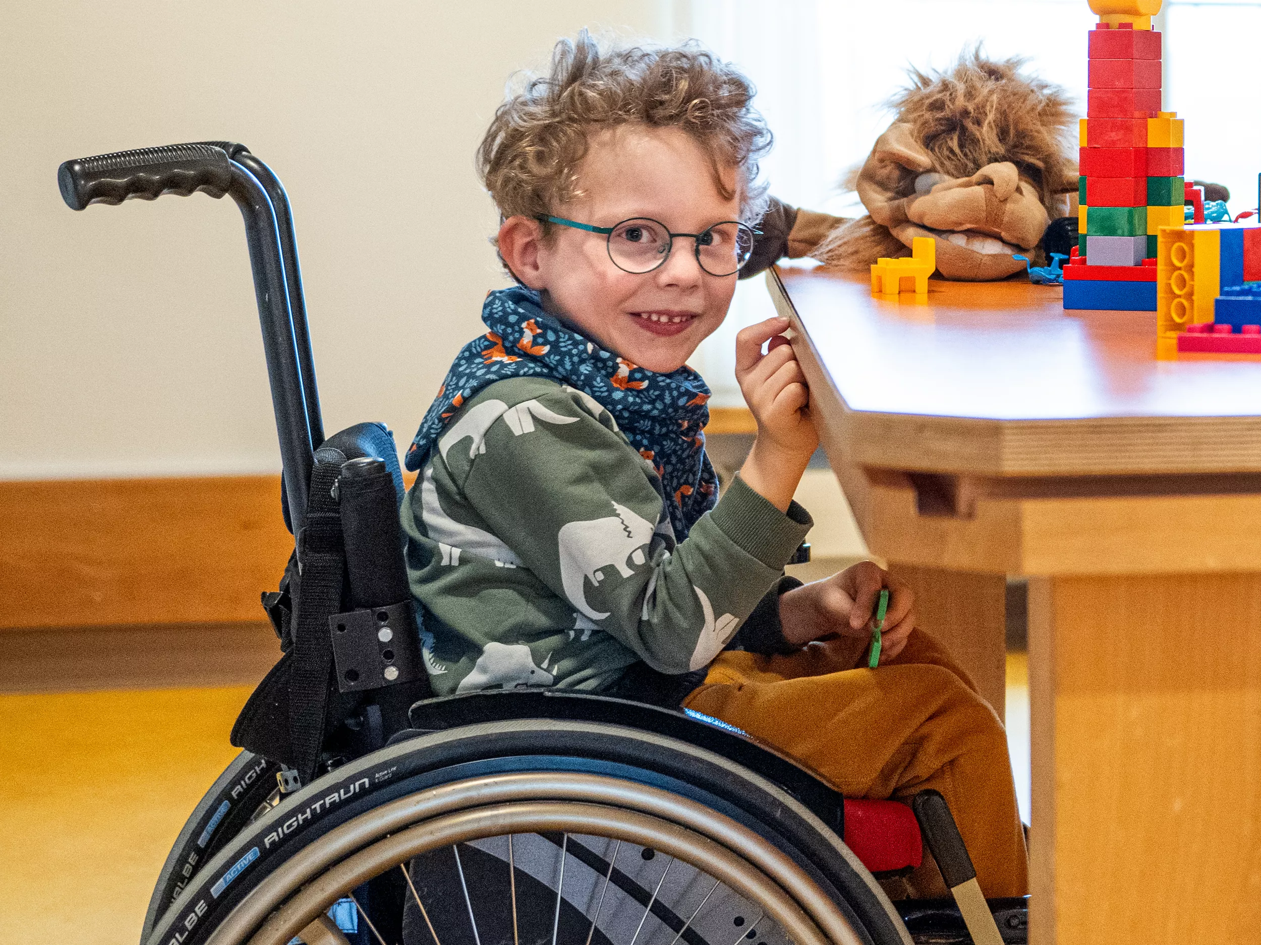 Ein junger Patient mit Brille sitzt in einem Rollstuhl und lächelt in die Kamera. Auf dem Tisch neben ihm liegt ein Kuscheltier und Spielzeug.