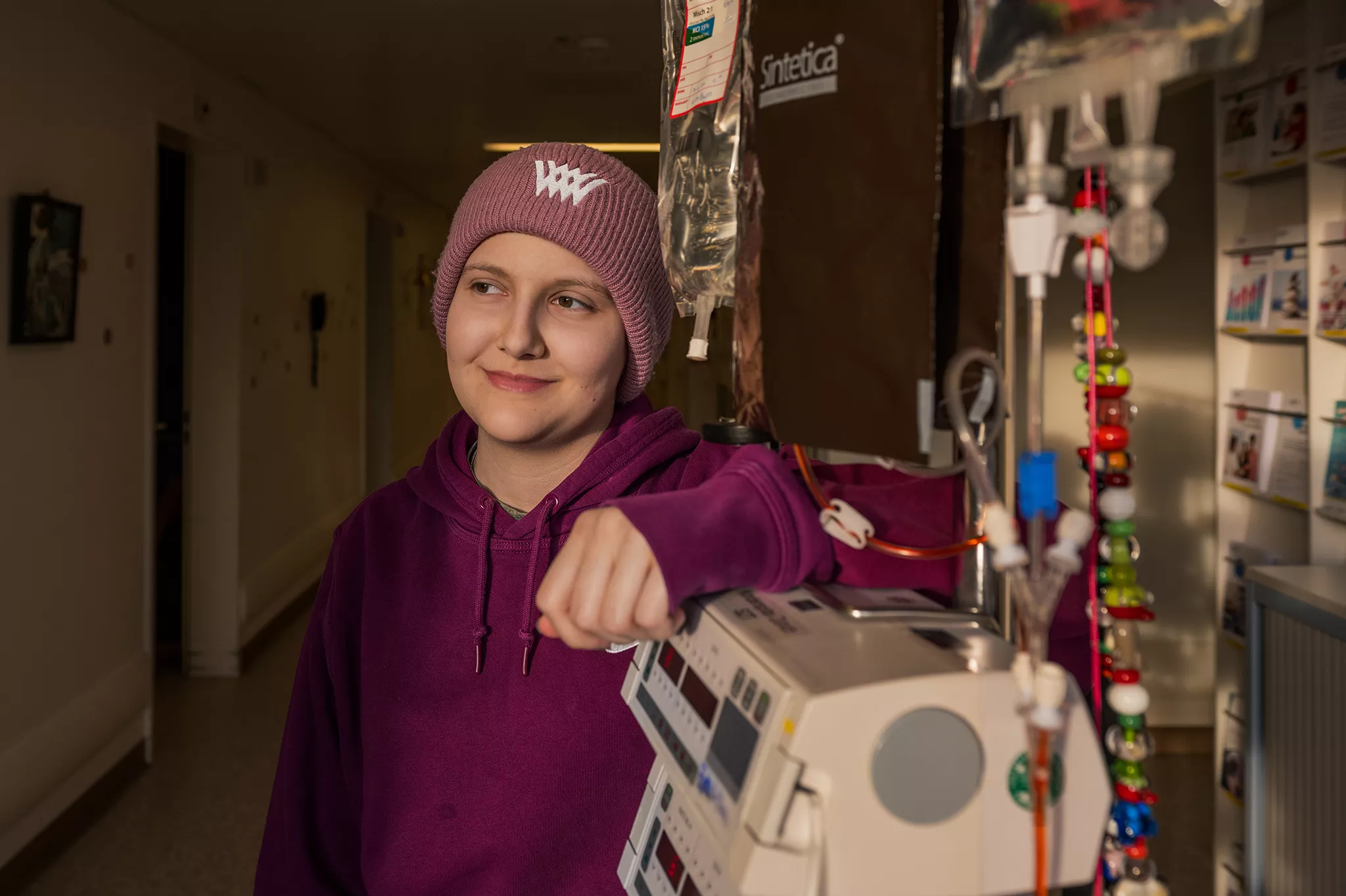 Eine Onkologiepatientin trägt eine Mütze und einen Pullover. Sie steht neben auf dem Gang neben einem "Chemoturm". 